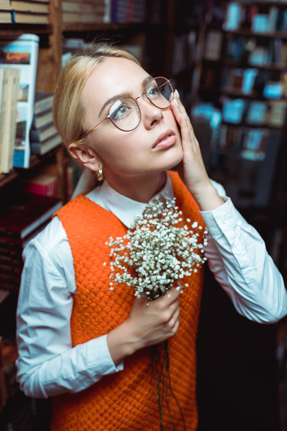 beautiful and blonde woman holding white flowers and looking up in library  - Foto, Imagem