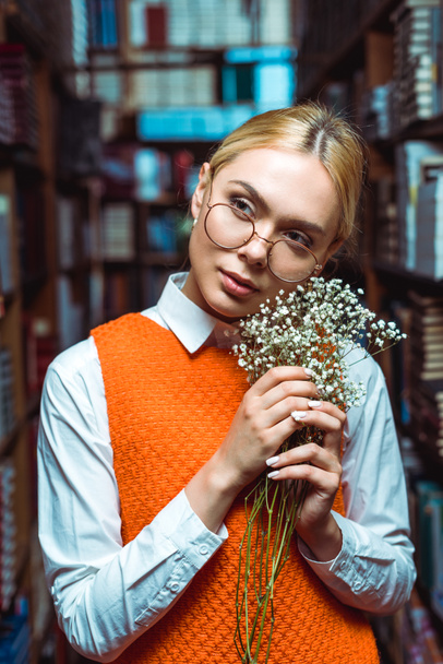 beautiful woman in glasses holding white flowers and looking away in library  - Zdjęcie, obraz