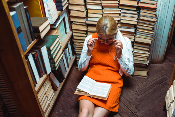 beautiful woman in orange dress sitting on floor and reading book in library - Foto, afbeelding