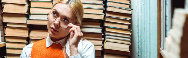 Panoramaaufnahme einer blonden und attraktiven Frau mit Brille, die in der Bibliothek wegschaut  - Foto, Bild