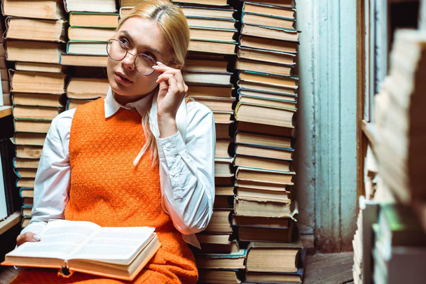  Blonde en aantrekkelijke vrouw in glazen houden boek en kijk weg in de bibliotheek  - Foto, afbeelding