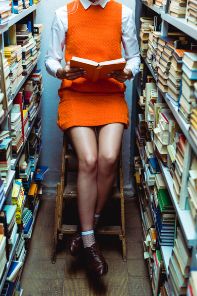 cropped view of woman in orange dress reading book and sitiing on ladder in library  - Fotografie, Obrázek