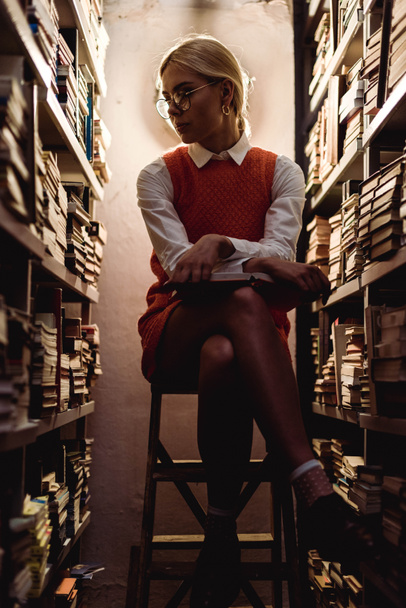 beautiful and blonde woman in orange dress sitting on ladder and holding book in library  - Photo, Image
