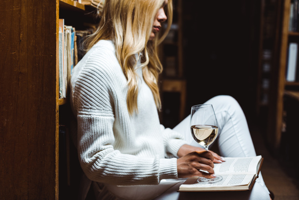 vista lateral de la mujer rubia leyendo libro y sosteniendo vino de cristal en la biblioteca
  - Foto, Imagen