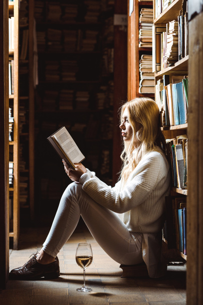 side view of blonde woman reading book and sitting on floor in library  - Fotó, kép