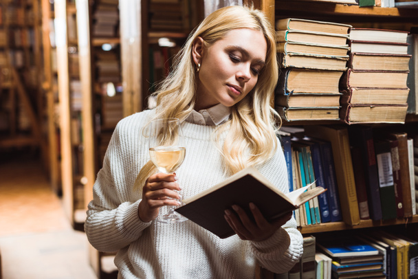 mujer rubia y bonita leyendo libro y sosteniendo copa de vino en la biblioteca
  - Foto, Imagen