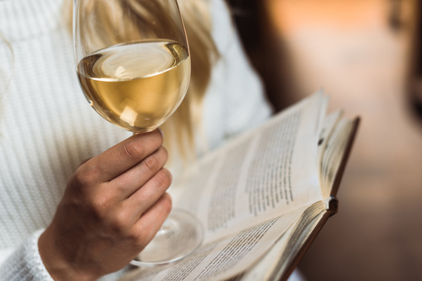 περικομμένη θέα της νεαρής ενήλικης γυναίκας που κρατά γυαλί κρασιού και βιβλίο  - Φωτογραφία, εικόνα