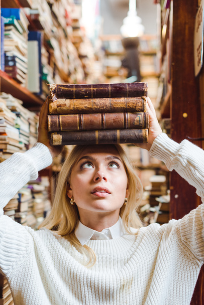 ξανθιά και όμορφη γυναίκα κρατώντας βιβλία και κοιτάζοντας πάνω στη βιβλιοθήκη  - Φωτογραφία, εικόνα