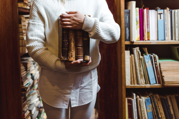 partial view of woman in white sweater holding books in library  - Foto, immagini