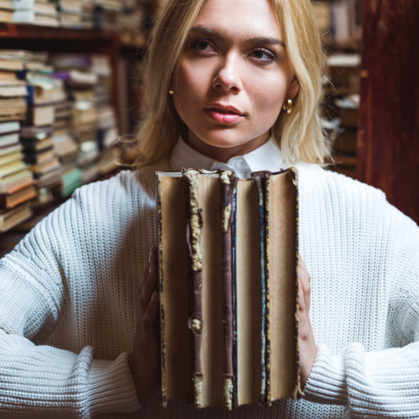 ξανθιά και όμορφη γυναίκα κρατώντας βιβλία και κοιτάζοντας μακριά στη βιβλιοθήκη  - Φωτογραφία, εικόνα