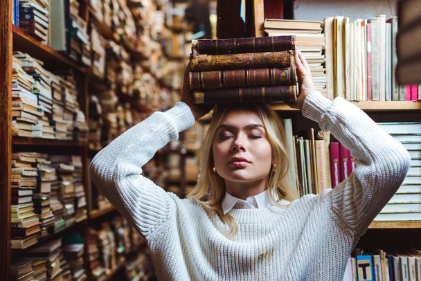 blonde and attractive woman with closed eyes holding books in library  - 写真・画像