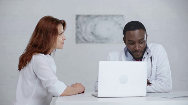 δύο πολυπολιτισμικούς γιατρούς που μιλούν και χαμογελούν ενώ κάθονται κοντά στο laptop  - Πλάνα, βίντεο