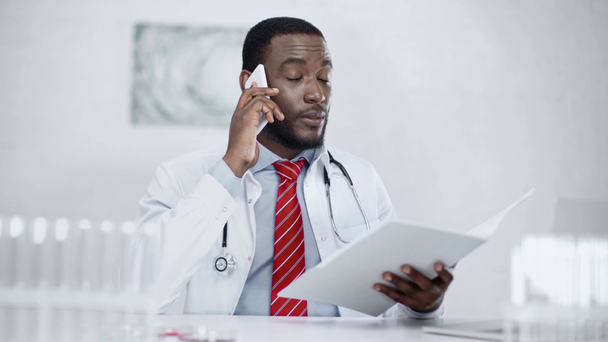 médico afroamericano serio hablando en el teléfono inteligente y la celebración de la carpeta de papel - Imágenes, Vídeo