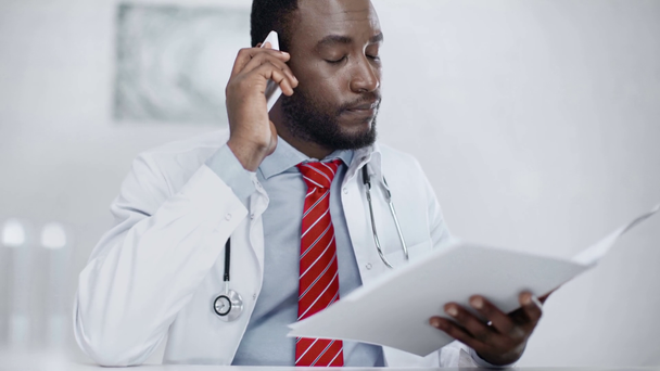 σοβαρός Αφρικανός Αμερικανός γιατρός κοιτάζοντας τα έγγραφα στο φάκελο, ενώ κάθονται στο χώρο εργασίας και μιλούν για smartphone  - Πλάνα, βίντεο