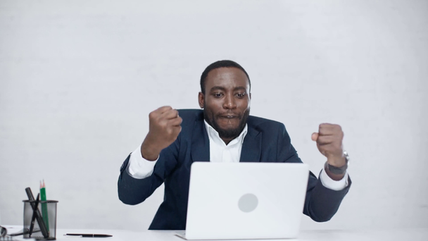 ενθουσιασμένος Αφρικανός Αμερικανός επιχειρηματίας χρησιμοποιώντας το laptop, δείχνοντας νικητή χειρονομία και χαμογελά στην κάμερα - Πλάνα, βίντεο