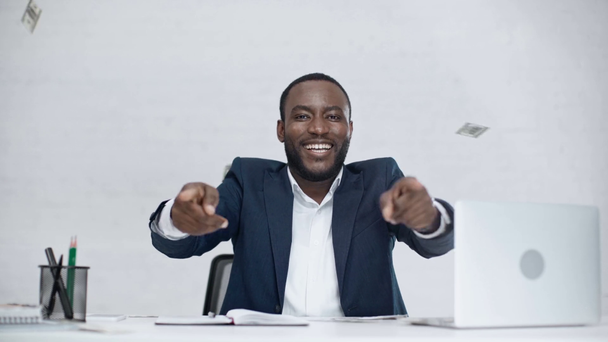glücklicher afrikanisch-amerikanischer Geschäftsmann wirft Dollarscheine hoch und lächelt in die Kamera, während er am Arbeitsplatz im Büro sitzt - Filmmaterial, Video