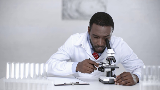 注意深いアフリカ系アメリカ人の科学者が顕微鏡で分析し、クリップボードに診断を書き込む - 映像、動画