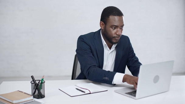 dissatisfied african american businessman using laptop and writing in notebook while sitting at workplace in office - Filmmaterial, Video