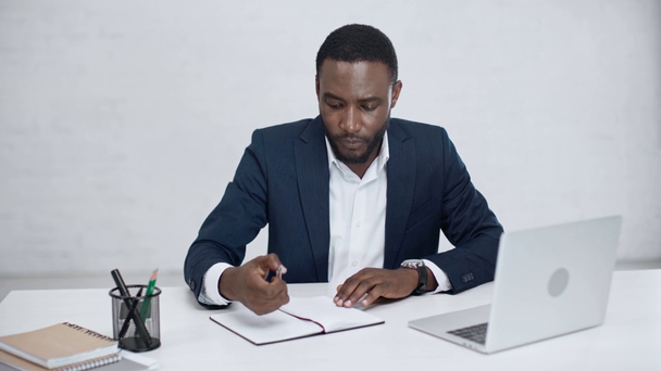 uważny African American biznesmen piśmie w notebooku i przy użyciu laptopa, siedząc przy biurku w biurze - Materiał filmowy, wideo
