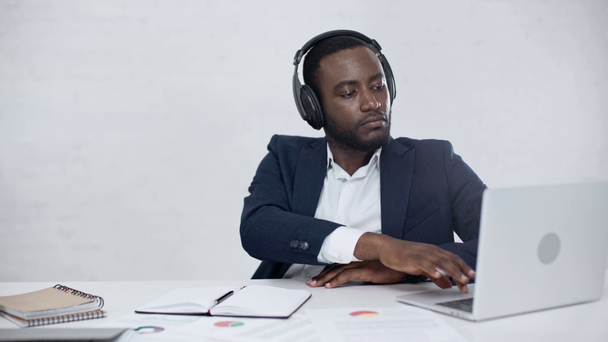 empresario africano positivo escuchando música en auriculares mientras está sentado en el lugar de trabajo cerca de la computadora portátil
 - Imágenes, Vídeo