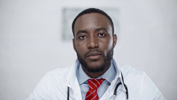 przystojny, wesoły African American lekarz uśmiechnięty i patrząc na aparat fotograficzny - Materiał filmowy, wideo