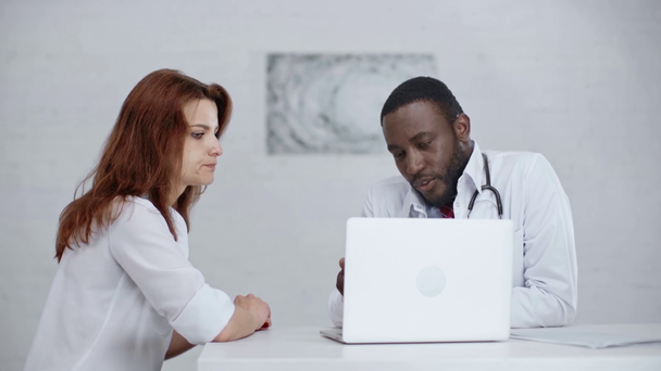 ラップトップの近くに座っている間、深刻なアフリカ系アメリカ人の医者が同僚と話す  - 映像、動画
