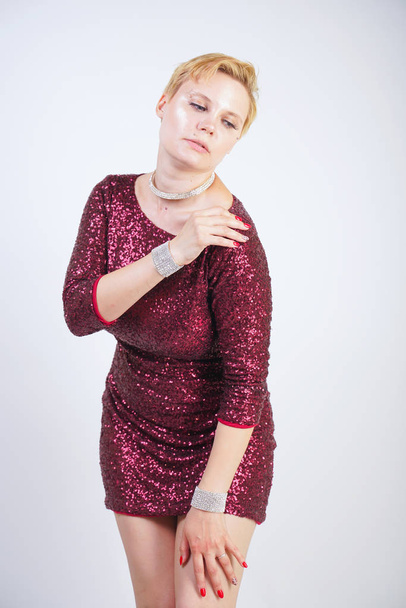 cute caucasian curvy girl with short blonde hair and plus size body wearing beautiful elegant cherry color dress with sequins and posing on a white background in the Studio alone. woman in party cloth - Photo, Image