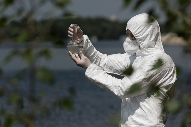 保護衣装、人工呼吸器、水サンプル付きフラスコを保持するゴーグルの水検査官の選択的な焦点 - 写真・画像
