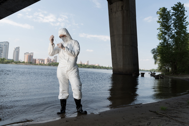 volledige lengte weergave van water inspecteur in beschermende kostuum en gasmasker nemen watermonster op rivier - Foto, afbeelding