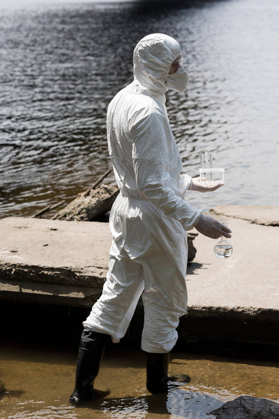 side view of water inspector in protective costume and respirator holding test tubes and flask with water samples at river - Fotografie, Obrázek