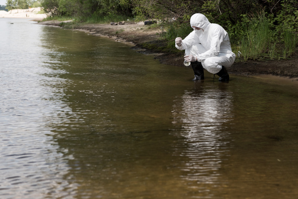 water inspector in protective costume, latex gloves and respirator taking water sample at river - Photo, Image
