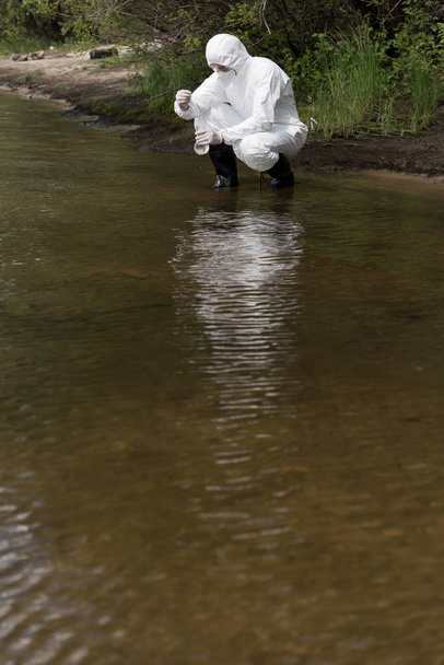 water inspector in protective costume, latex gloves and respirator taking water sample at river - Photo, Image