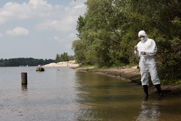 full length view of water inspector in protective costume, latex gloves and respirator holding flask with water sample at river - Foto, Imagem