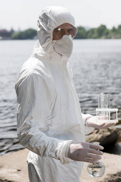 water inspecteur in beschermende kostuum, latex handschoenen en gasmasker houder kolf en reageerbuisjes met watermonsters aan rivier - Foto, afbeelding