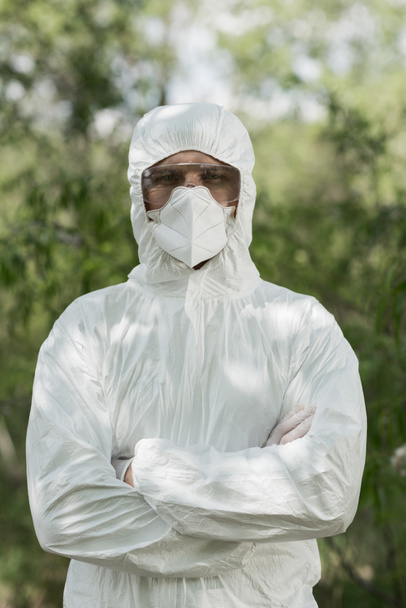 front view of ecologist in protective costume and respirator with crossed arms in forest - Photo, Image