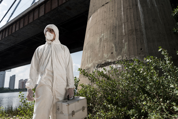 ecoloog in beschermend kostuum met inspectie Kit staande in de buurt van Bridge - Foto, afbeelding