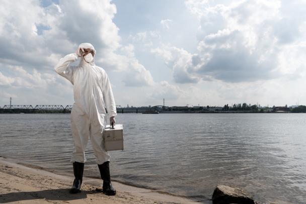 vista completa del inspector de agua en traje de protección que sostiene el kit de inspección en la costa del río
 - Foto, imagen