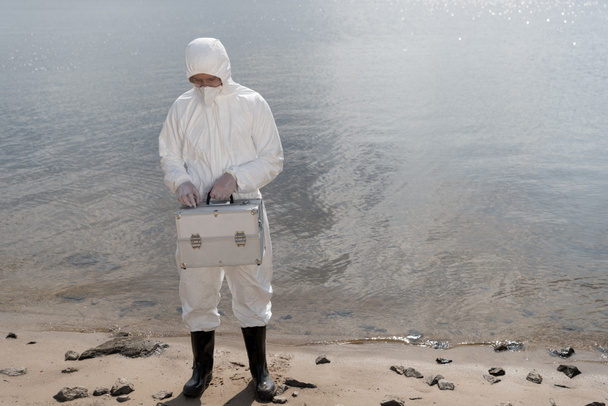 volledige lengte weergave van water inspecteur in beschermende kostuum Holding inspectie Kit aan rivier kust - Foto, afbeelding
