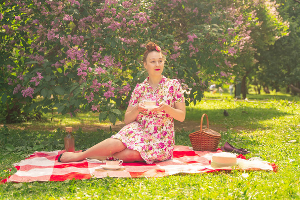 sweetheart charming pinup girl in a summer dress on a checkered blanket in the Park near the bushes of lilac enjoys life and leisure alone. pretty happy caucasian woman on the grass. - Φωτογραφία, εικόνα