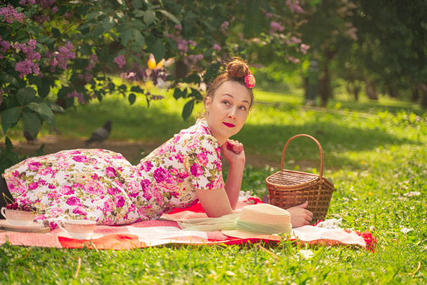 Kochanie uroczy Pinup dziewczyna w letniej sukni na koc w kratkę w parku w pobliżu krzewów bzu cieszy się życiem i wypoczynku sam. całkiem szczęśliwy kaukaski kobieta na trawie. - Zdjęcie, obraz