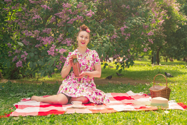 corazón encantadora pinup girl en un vestido de verano en una manta a cuadros en el parque cerca de los arbustos de lila disfruta de la vida y el ocio solo. bastante feliz mujer caucásica en la hierba
. - Foto, imagen