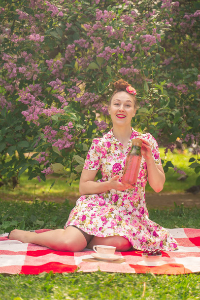 corazón encantadora pinup girl en un vestido de verano en una manta a cuadros en el parque cerca de los arbustos de lila disfruta de la vida y el ocio solo. bastante feliz mujer caucásica en la hierba
. - Foto, Imagen