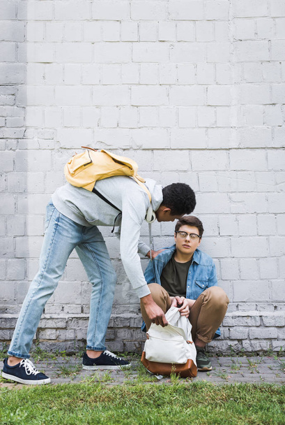 aggressive african american boy taking backpack from frightened boy in glasses  - Фото, изображение