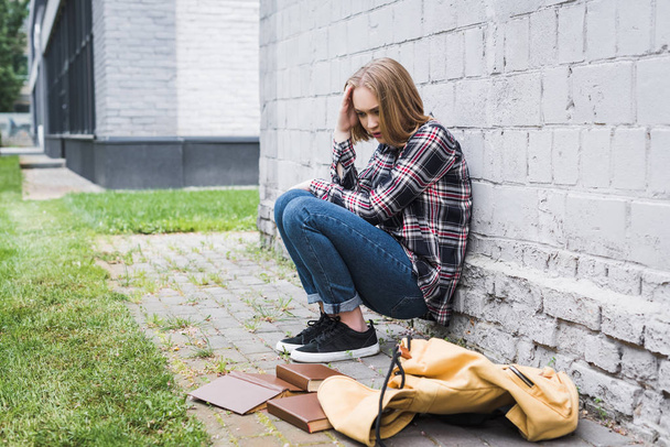 sad and blonde teen in shirt and jeans sitting near wall among books  - Φωτογραφία, εικόνα