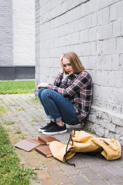 disappointed and blonde teen in shirt and jeans sitting near wall among books  - 写真・画像