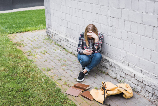 disappointed and blonde teen in shirt and jeans sitting near wall and holding smartphone  - Foto, Imagem