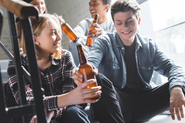 smiling teenagers sitting on stairs, drinking beer and talking  - Φωτογραφία, εικόνα