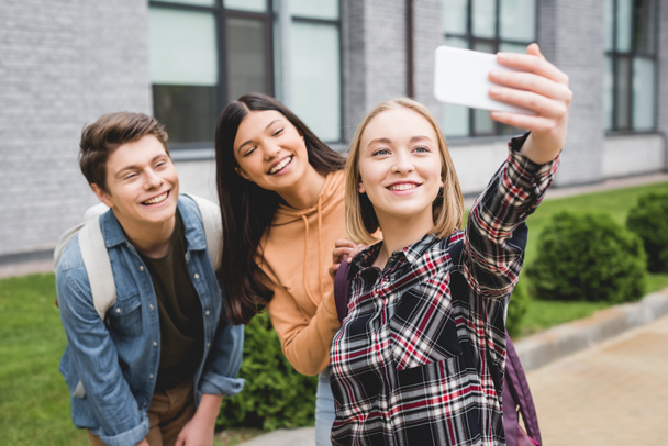 cheerful teenagers holding smartphone, taking selfie and smiling outside - Фото, зображення
