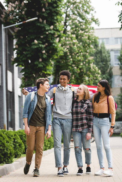 Lächelnde und glückliche Teenager, die eine amerikanische Fahne halten und miteinander reden  - Foto, Bild
