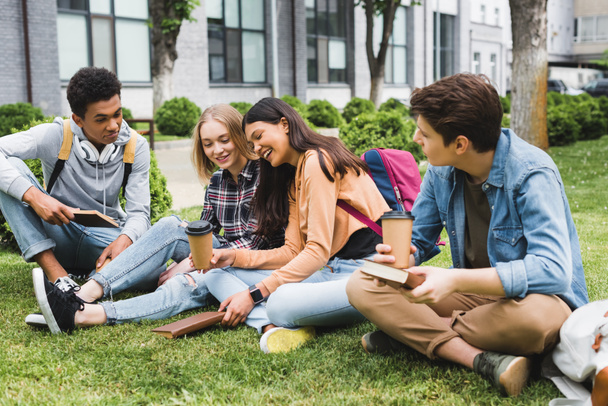 adolescents souriants assis sur l'herbe, parlant, tenant des livres et des tasses en papier
  - Photo, image
